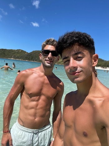 Dos hombres jóvenes sonrientes, con torso descubierto, posan para una selfie en agua turquesa; uno usa gafas de sol, el otro guiña un ojo, con colinas verdes al fondo