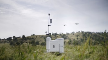 Un dispositivo blanco de tecnología Rainmaker con una antena en un campo verde, con colinas arboladas y dos drones volando bajo un cielo nublado