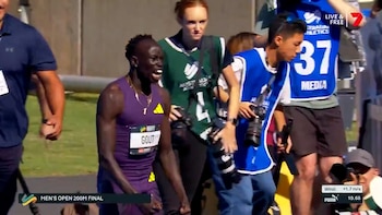Atleta Gout Gout, hombre de piel oscura con camiseta morada y expresión eufórica, celebra en pista. Rodeado de fotógrafos y personal de medios