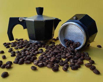 Una cafetera moka de metal y negra, una parte en posición vertical y la otra tumbada, rodeadas de granos de café sobre un fondo amarillo