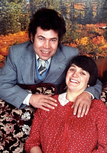 Hombre y mujer sonriendo. Él viste traje gris, chaleco azul y corbata. Ella lleva un vestido rojo con puntos blancos y cuello blanco. Fondo con arbustos