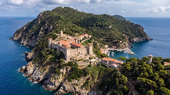 Una vista aérea de una isla montañosa con vegetación, edificaciones fortificadas de piedra y un puerto pequeño con barcos.