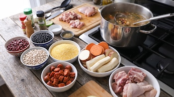 Ingredientes para un guiso como frijoles, maíz, chorizo, carne cruda, yuca y camote en tazones sobre una mesa de madera, con una olla de estofado en la estufa.