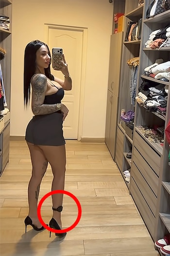 Una mujer frente a un espejo, con una tobillera electrónica en el tobillo derecho, posando en un vestidor con ropa y accesorios