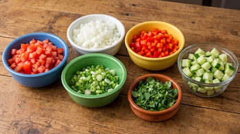 Seis bowls de colores y transparentes con tomates, cebolla blanca, pimentón rojo, cebolla larga, cilantro y pepino picados en cubos sobre una mesa de madera.
