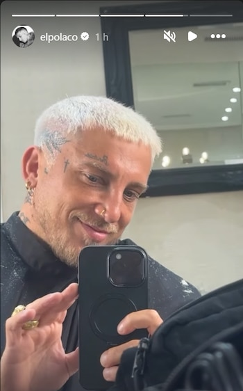 Hombre con cabello corto platinado y tatuajes faciales toma una foto frente a un espejo sosteniendo un teléfono celular negro