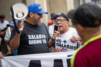 Primer plano de dos hombres adultos participando en una protesta. Uno sostiene un megáfono y el otro habla por un micrófono, mientras muestra un cartel