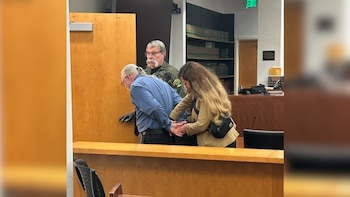 Mitchell Gaff, un hombre canoso de coleta y gafas, es esposado por una mujer en una sala de tribunal, mientras un oficial uniformado observa detrás. Se ven estanterías y paneles de madera