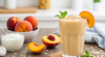 Un vaso de licuado de durazno con avena y menta, rodeado de duraznos enteros y cortados, avena, y un recipiente de yogur en una mesa de madera.