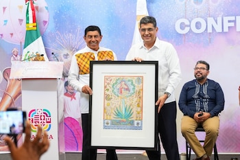 Dos hombres sonrientes sostienen un cuadro con una ilustración colorida, flanqueados por un podio con el logo de Guelaguetza y una bandera de México