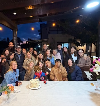 Un grupo numeroso de personas posa detrás de una mesa con un pastel, flores y copas en una galería iluminada con guirnaldas