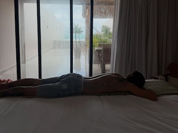 Un hombre con tatuajes en la espalda descansa boca abajo en una cama frente a un ventanal por el que se ve una terraza y el mar