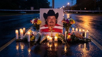 Fotografía de un memorial con una imagen de un hombre con sombrero, rodeado de velas encendidas y ramos de flores en una carretera mojada al anochecer.