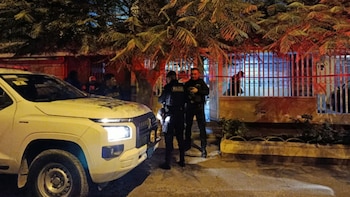 Vista nocturna de dos agentes de policía y un vehículo policial blanco frente a una vivienda con árboles y cinta de seguridad