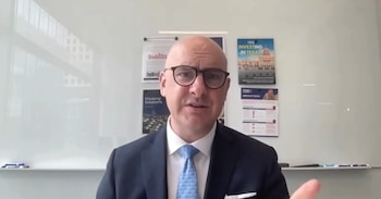 Hombre calvo con gafas y traje oscuro, Jeremy Mazur, habla ante una pared blanca con gráficos y pósteres informativos