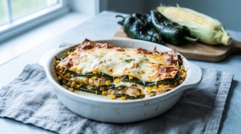 Una fuente de cerámica blanca con lasaña poblana de pollo, elote y queso gratinado, sobre un paño de lino gris. Al fondo, chiles poblanos y una mazorca de maíz.