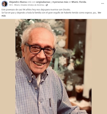 Hombre mayor de pelo canoso y gafas sonriendo ampliamente, vestido con una camisa a cuadros azul y blanca; se aprecia un árbol de Navidad borroso al fondo