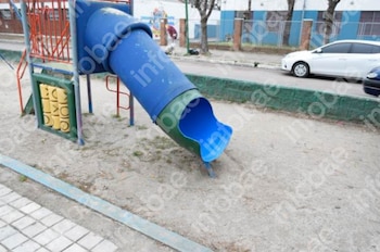 Tobogán de tubo azul y verde en un parque infantil de tierra. Se observan barras rojas, panel de juego amarillo, acera y coches en la calle