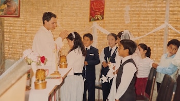 Un sacerdote con vestidura blanca bendice a una niña junto a un grupo de niños vestidos formalmente frente a una pared de esteras