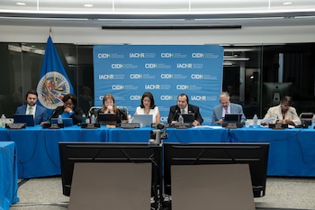 Seis personas sentadas en una mesa azul con laptops, micrófonos, botellas de agua y placas de identificación. Una bandera y un panel con logos de la CIDH al fondo