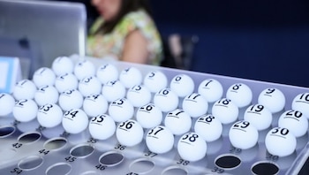 Un arreglo de bolillos blancos numerados en una bandeja transparente. Al fondo se observa una persona y una pantalla de ordenador borrosas