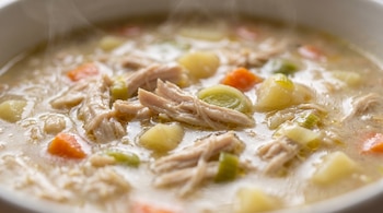 Primer plano de un tazón de sopa humeante con pollo desmenuzado, trozos de papa, zanahoria y puerro en un caldo cremoso