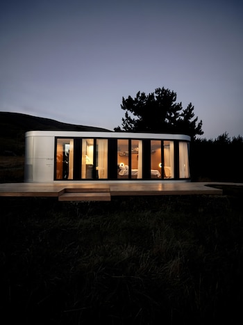 Una estructura de metal con amplios ventanales iluminados por dentro se alza sobre una plataforma de madera en el campo al anochecer