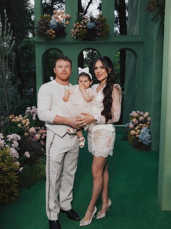 Hombre, mujer y bebé posan de pie en un jardín con césped verde artificial y un muro decorado con arcos y arreglos florales de colores