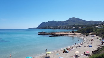 Playa con personas sobre la arena, sombrillas de colores, agua turquesa y montañas en el fondo bajo un cielo despejado.