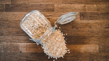 Un frasco de vidrio tumbado con la tapa abierta y copos de avena esparcidos sobre una tabla de madera.