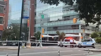Calle urbana con edificios, dos vehículos policiales con luces encendidas, cinta policial, dos agentes de policía, señales de tráfico y semáforos