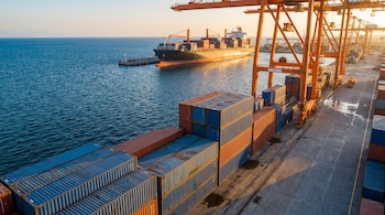 Filas de contenedores de carga azules y naranjas en un muelle portuario con grúas industriales y un buque de carga en el mar.
