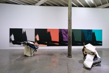 Galería de arte con grandes paneles abstractos de colores vibrantes en la pared, dos esculturas de metal blanco aplastado en el suelo y una columna oxidada