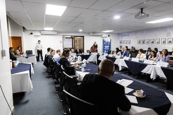 Una sala de conferencias con mesas en forma de U, personas sentadas, proyector de techo, botellas de agua, tazas, folletos y una pancarta de Guatemala