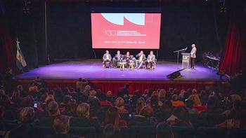 Escenario de auditorio con un panel de cinco personas sentadas, un orador de pie en un podio, una pantalla grande y una audiencia