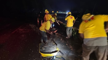 Cinco trabajadores con camisas amarillas y gorras realizan labores de limpieza en una carretera oscura con conos y señales viales. Uno vacía tierra con una pala en una carretilla