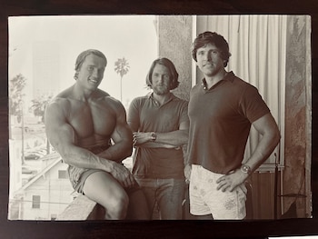 Fotografía en blanco y negro de tres hombres. Un hombre musculoso sin camisa, un hombre con barba y un hombre con polo. Edificios y palmeras al fondo