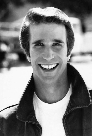 Retrato en blanco y negro de Henry Winkler sonriendo, luciendo cabello peinado hacia atrás, camiseta blanca y chaqueta de cuero abierta