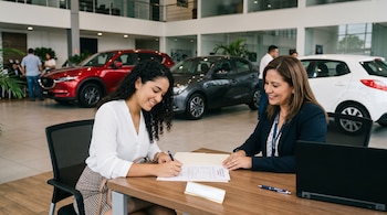 Una mujer firma un papel sentada en un escritorio junto a otra persona frente a una computadora, con tres automóviles de fondo.
