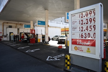 Primer plano de un cartel de precios en una estación Shell. Se ven los precios de Super, V-Power Nafta, Diesel y GNC. Al fondo hay vehículos y personal