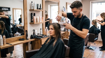Un peluquero con delantal negro corta con tijeras el cabello húmedo de una mujer sentada en una peluquería.