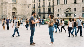 Pareja de pie conversando en una plaza abierta con edificios de piedra antiguos y una fuente al fondo. Numerosos peatones transitan alrededor.