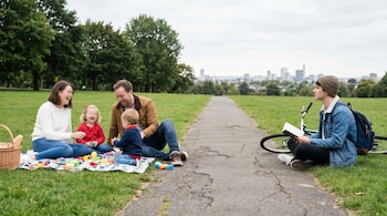 Familia de cuatro en una manta de picnic con juguetes y un joven adulto leyendo un libro, sentados sobre césped junto a un sendero y una bicicleta. Fondo con árboles y edificios.