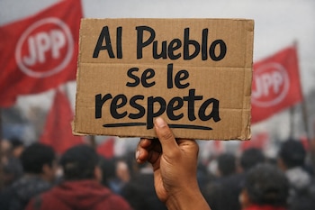 Primer plano de una mano sosteniendo un cartel de cartón con el mensaje 'Al Pueblo se le respeta' escrito en negro. Al fondo, banderas rojas de JPP y manifestantes.