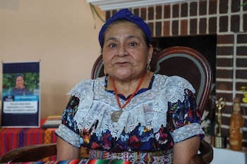 Mujer sentada en una silla, viste tocado azul, blusa bordada con encaje, collar de cuentas rojas y medallón plateado. Fondo con afiche y pared de ladrillo