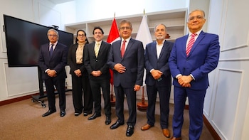 Seis personas en traje formal posan de pie en una oficina frente a una bandera peruana y un televisor de pantalla plana