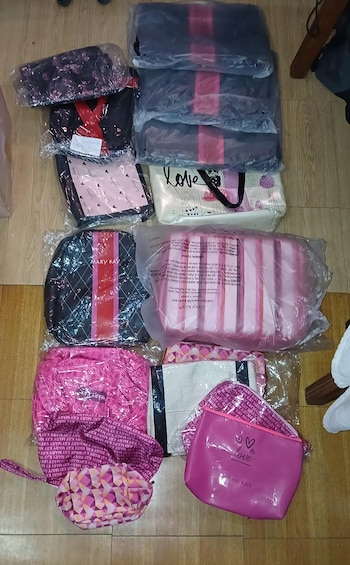 Diversos bolsos, mochilas y cosmetiqueras de colores rosa y negro, algunos en bolsas plásticas, organizados sobre un piso de madera