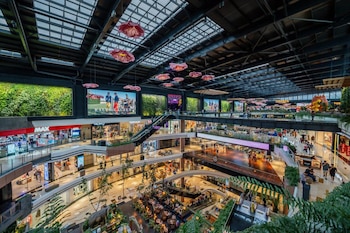 Vista interior de un centro comercial de varios niveles con tiendas, escaleras mecánicas, vegetación y flores colgantes bajo un tragaluz