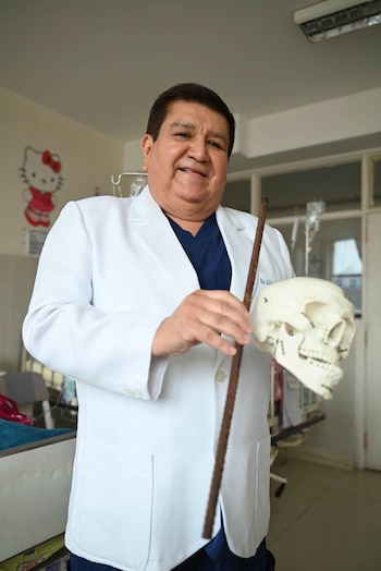 Un médico con bata blanca y uniforme azul sostiene una varilla de construcción y un modelo de cráneo. Al fondo se observa una habitación de hospital con un dibujo de Hello Kitty