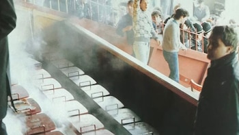 Humo asciende de asientos de estadio, algunos dañados, en primer plano. Personas evacúan por una rampa en el fondo y una figura en el lateral derecho